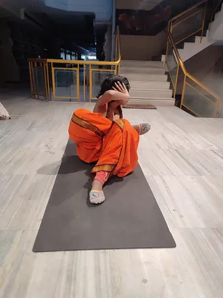 A single person sits on a dark grey yoga mat on a light marble floor inside a building with gold-railed glass balustrades and a white staircase behind. The person wears an orange garment with a gold-and-maroon border, has a rudraksha mala around the neck, and wears grey socks with a small colourful logo. One hand is raised to the head in a seated yoga pose. The figure shows distinctive long dark hair and a bare upper body with red beads, which are cues for a person, but the face and upper body are partly obscured, so the identity is uncertain.