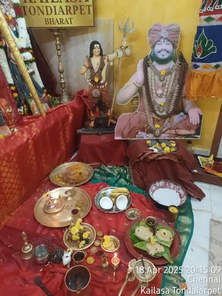 Photograph of a worship set-up inside a temple room with a yellow wall and a sign reading KAILASA TONDAIARPET BHARAT. Two large life-size cutout portraits stand on a draped red and gold table: on the right a seated figure in a pink turban with a rudraksha mala, white and gold necklaces, and a raised hand, and on the left a standing figure holding a trident, wearing a rudraksha-beaded vest and dhoti. Both are printed portraits, not physical people. In front, a red cloth covers the floor with brass plates holding coconut halves, bananas, steamed rice cakes on leaves, yellow flowers, coins, a brass kalasha-style vessel, a small oil lamp, a bell and a copper pot of water. A timestamp reads 18 Apr 2025 20:15:09 Chennai. No physical people are visible.
