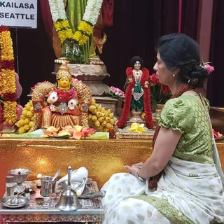 A woman in a green and white sari sits cross-legged before a shrine decorated with marigold and rose garlands. She wears a rudraksha mala and wrist bangles and has a flower in her hair. The shrine holds a gold-and-red deity murti flanked by bananas and rose petals, a small standing figure in red garland and green dhoti with a tall black headpiece, a silver handbell, oil lamps, a brass pot and a silver dish. The scene looks like a puja at the KAILASA Seattle centre.