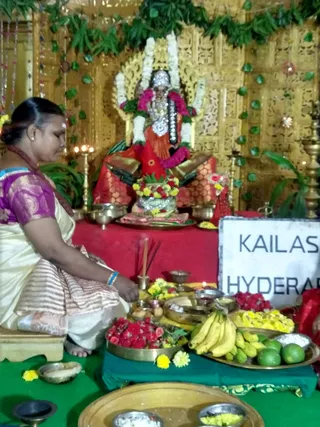 A woman in a cream and maroon-striped silk sari with a purple blouse sits cross-legged at a puja setup, a red-draped table before a golden lattice backdrop, and performs worship before a decorated deity murti of Lakshmi, adorned with white and pink garlands and a silver crown. Brass lamps, incense sticks, brass plates of red flowers, yellow marigolds, bananas, limes and coconut shavings, and a kalasha with flowers surround the altar. A white sign reads KAILASA HYDERABAD, consistent with the Varalaxmi Vratham seva.