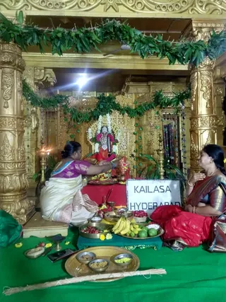 A photograph of a Varalakshmi puja at a gilded shrine. A deity murti of Lakshmi, adorned with white and pink garlands, sits at the centre within a gold lattice backdrop hung with green leaf garlands. Two women sit on a green cloth before a red-draped table: one in a magenta blouse and cream sari offers flowers toward the deity, and the other in a red silk sari sits with hands folded in prayer. Offerings include bananas, limes, fruit, betel, coconut-like pieces, brass bowls of rice and yellow grains, and a brass lamp. A sign reads KAILASA HYDERABAD. Two physical people are visible.