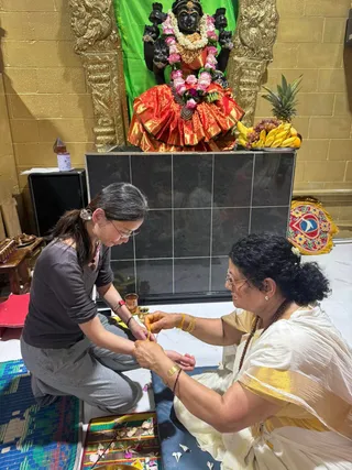 Indoor temple scene in front of a black deity murti with a green backdrop, decorated with pink roses, white jasmine garland and a red-and-gold silk skirt; a fruit offering of bananas, grapes and a pineapple sits to the right. Two women sit on the floor: a woman in a grey top and grey trousers, wearing glasses, ties a yellow thread on the wrist of an older woman in a white and gold-bordered Kerala-style saree, with a rudraksha mala and gold bangles, who holds the thread. Petals lie on a striped mat and a brass vessel stands nearby. No a person candidate is visible.