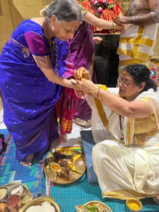 A photograph of a ritual in a temple-like room with a yellow-tiled wall. A woman in a royal blue silk sari with gold motifs and a rudraksha mala stands at left, bent forward while a woman in a white sari with a gold border sits cross-legged at right, tying or fastening a thread around the standing woman's wrist. A third person in a white dhoti with gold border stands behind them, and another woman in a magenta and gold sari kneels behind. In front, a brass tray holds brass oil lamps with lit flames, a ripe banana, incense sticks, a small bowl of kumkum, and a bowl of yellow turmeric. Brass plates of food, including idli and rice, sit on the blue and green mat at lower left and lower right. A red and gold deity murti is partly visible at the top.
