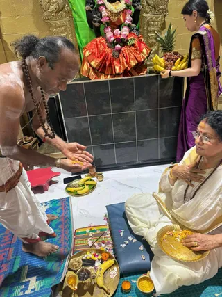 Photo of a puja inside a temple shrine. A shirtless man with a topknot and rudraksha malas, wearing a white dhoti, stands at left holding turmeric or saffron in his palm over a brass plate of bananas. Seated at right on a blue cushion, a woman in a white sari with a gold border and glasses, with a red bindi, holds a gold plate of flower petals and turmeric. Behind them a woman in a purple sari holds a tray of bananas, pineapple and grapes before a deity murti draped in a red and gold silk garment, decorated with pink roses and a garland. Brass oil lamps, a kalasha-style vessel, incense and marigold petals lie on the floor mat and tiles. Black and gold temple walls frame the shrine.