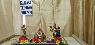 A photograph of a shrine table set against gold draped curtains, with a white sign reading KAILASA TRINIDAD & TOBAGO in black capitals taped above. On the altar stand two copper-coloured deity murtis: a small figure on the left holding a bunch of flowers, and a taller seated figure on the right with a crown, holding flowers and a small figure in its lap, wearing a green cloth. A tiered copper object, resembling a shivalingam or yantra base, sits between them. Red, yellow and blue flowers and yellow plumeria blossoms decorate the platform. No people are visible.
