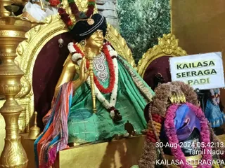 A golden statue seated on a gold-framed throne with maroon cushions, dressed in a green garment with silver-threaded checks and a multicoloured striped shawl draped over one shoulder. The statue wears a black turban with a brooch, red and white flower garlands, a silver ornamental collar, and holds a golden staff in one raised hand. A brass bell and oil lamp stand nearby, and to the right a shivalinga-style object is decorated with purple, pink and red garlands. A white sign reads KAILASA SEERAGAPADI. A timestamp on the photo reads 30-Aug-2024 05:42:13 Tamil Nadu.