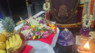 Photograph of a temple altar set up for worship. A heap of mixed flowers, including pink roses, white jasmine-type blossoms, purple and yellow blooms and green leaves, rests on a red cloth on a white-draped table. Beside it stand a pineapple, bananas and other fruit in a terracotta bowl. Two small metal deity statues are adorned with white flower garlands: one standing figure in a brown-and-gold garment at the back, and one on the right in a purple sari-style dress with a crown. A folded purple brocade cloth with gold trim sits at right, and a lit oil lamp glows in the lower right foreground. A dark brown-and-gold cloth backdrop with a printed deity image hangs behind the altar, with a garland of marigolds draped over it. No people are visible.