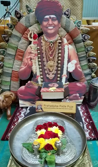 A large life-size cutout photograph of a smiling man in a maroon turban and maroon robe sits cross-legged on a throne-like seat with a beaded, striped backdrop. He holds a silver trident adorned with a red flower in one hand and a white flower in the other, wearing layered gold and bead necklaces and a white garland. In front of the cutout sits a book with a portrait on its cover and a yellow flower. A wooden elephant stands at the left edge. In the foreground, a silver tray holds red and yellow flowers and green leaves between two silver vessels. A yellow sign reads Pratyaksha Pada Puja, offering your gratitude at the divine lotus feet of swamiji. The cutout is printed, so no physical people are visible.