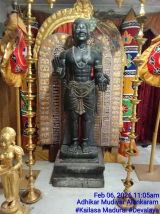 A dark standing deity murti, carved in black stone, stands on a stepped pedestal in front of a gold arched backdrop with mirror-work and a peacock-feather fan. The figure has a tilak on the forehead, a beaded necklace, a sash at the waist, and bare arms raised in a blessing gesture. Tall brass lamp stands with multiple tiers flank the murti, and a gilded female figure holding a lamp stands to the left. Red and multicoloured floral silk drapes and a kalasha-like finial frame the scene. Overlay text reads Feb 06, 2026 11:05am, Adhikar Mudivar Alankaram and #Kailasa Madurai #Devalaya. No people are visible.