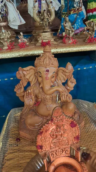 Close photo of a terracotta-coloured Ganesha murti seated on a woven mat before a blue silk backdrop. The four-armed elephant-headed deity wears an ornate crown, a red-and-white tilak on his forehead, and a beaded necklace. Red flowers are scattered around the figure. In front stands a copper-coloured kalasha or ritual vessel decorated with a repoussé deity, with red kumkum-like powder on it. Above on a gold-trimmed shelf are smaller brass and gold-toned deity figures, some in blue and white garments, with red flowers placed among them.