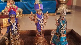 Photograph of three small wooden or metal murti figures displayed on a dark tray in a temple setting. The central figure is a multi-armed deity wearing a purple cloth, a tall pearl-studded crown with green top, rudraksha mala and layered white beaded necklaces, with kumkum marks on the forehead. Figures to the left and right wear purple and teal sarees with similar pearl crowns and necklaces. Behind them stand a blue cloth backdrop, brass vessels, a gold-toned pot and a small ganesha figure. Shiva lingam-like objects appear at the lower left. No people are visible.