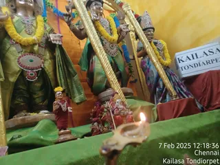 Photograph of an altar with several large painted deity murtis dressed in green and pink silk garments, adorned with yellow marigold garlands and jeweled ornaments. A small statue of a figure in a maroon robe with a golden crown and a Ganesha figure sit on a green cloth in front, with a lit oil lamp in the foreground. A white sign reads KAILASA TONDIARPET. A timestamp shows 7 Feb 2025 12:18:36 Chennai Kailasa Tondiorpet. No physical people are visible.