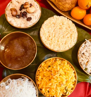 A top-down photograph of a ritual food offering arranged on a large green leaf over a red cloth. Small brass bowls hold white rice topped with cashews and raisins, plain white rice, a dark amber sweet preparation, bright yellow tempered rice with cumin seeds, and grated white coconut. A brass tray at the top right holds whole orange fruits and a gourd. No people are visible; the arrangement is a prasadam-style food offering for a puja.