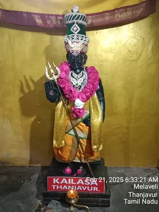 A photograph of a black stone deity murti standing inside a shrine against a gold-painted wall with a maroon and gold cloth canopy above. The figure wears a tall green and silver crown with jeweled bands, a pink flower garland and a jeweled necklace, and a gold brocade shawl over an orange dhoti. It holds a silver trident in one hand. Flower petals lie at its base and a brass oil lamp burns in front of the red-and-white pedestal sign. No people are visible.