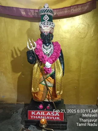 A dark stone or painted statue of a standing deity murti, with a tall green and silver ornate crown, a trident held in its raised hand, a pink and white flower garland, a jewelled silver necklace, and a gold-bordered orange dhoti with a gold shawl. A brass oil lamp burns in the foreground before a red nameplate on the pedestal, against a gold wall with a maroon cloth canopy above. Timestamp overlay reads Feb 21, 2025 6:33:11 AM, Melaveli, Thanjavur, Tamil Nadu. No people are visible.