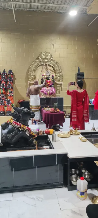 A photograph inside a temple-style hall with yellow-painted block walls and a gold ornate arch framing a black deity murti decorated with garlands and a pink-and-gold sari. A bald priest with a bare chest and white dhoti with gold border stands at left, raising a lit oil lamp toward the deity. Near the centre a woman in a red salwar kameez with red-and-gold dupatta stands behind a maroon-draped table holding fruit, a pineapple and a lamp, and photographs the scene with her phone. In the foreground a black Nandi bull statue wears a red and white garland, and brass plates, cups and a bell sit on a white counter. Shelves at left hold rows of small orange and green garlanded deity figures.