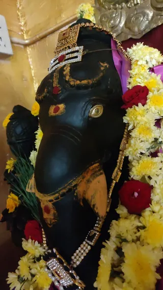 Close-up photograph of a black stone Ganesha murti face, decorated with yellow and white chrysanthemum garlands, red roses, and green grass. The deity wears a gold crown with red stone and white rhinestone embellishments, gold-rimmed eyes, red kumkum marks on the forehead and trunk, and a jewelled necklace with pearls and rhinestones. A purple cloth is draped behind the head. No people are visible.