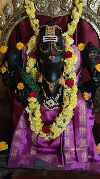 A black stone Ganesha murti with an elephant head is seated in a shrine, dressed in a magenta silk cloth with gold zari borders. It wears a gold and white-stone crown ornament with a red centre stone, a matching pearl and gem necklace, and yellow chrysanthemum garlands with red rose accents. Yellow flowers are scattered around the image, and a green grass blade rests against one arm. Behind it is a deep maroon drape and an ornate gilded arch. No people are visible.