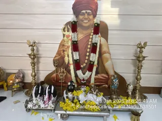 A photograph of a devotional shrine set up for Pada Pooja. Centrally, a large printed cutout portrait shows a smiling man in a red-orange turban, saffron-orange robe, rudraksha-style bead necklace and garland of red, white and green flowers, holding a trident-topped staff. In front, on a silver tray, stand a black shiva-lingam-like form draped in white cloth with yellow marigolds, and a pair of black sandals (paduka) with white flowers on a second tray. Brass oil lamps with bird-shaped tops flank the shrine, and a small brass vessel and yellow flower petals lie on the white table. No living people are visible.