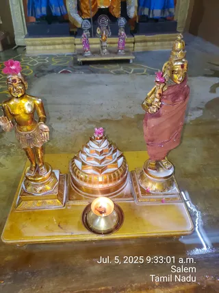 A photograph of a small shrine arrangement on a gold-coloured base in a hall with a patterned floor. Two gilded standing figures flank a white and gold stepped sri yantra meru at centre, with a small brass oil lamp burning in front. The figure on the left is bare-chested with a short gold dhoti; the figure on the right wears a red-brown draped cloth and a gold crown. Both have pink flowers tucked near their heads and red kumkum marks on their foreheads. Behind them stand smaller painted figures in pink and blue robes and a deity murti, with blue-and-gold drapes above. A timestamp reads Jul. 5, 2025 9:33:01 a.m., Salem, Tamil Nadu.