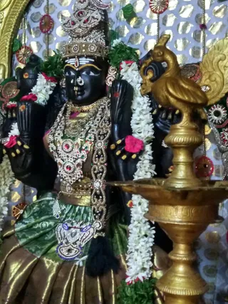 A black stone deity murti stands in a shrine decorated with white jasmine garlands, red flowers and a gold-and-silver foil backdrop with red and gold ornaments. The murti wears a silver-jewelled crown, a tilak on the forehead, a nose ring, heavy gold and gem-studded necklaces, and a green and gold silk garment with a jewelled waist belt. A tall brass oil lamp with a bird-shaped top stands in the foreground on the right. No people are visible.