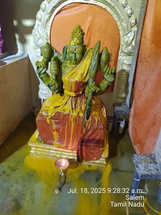 A photograph of a seated deity murti in a shrine, its green-painted body smeared with yellow turmeric paste and draped in a red-orange cloth with a yellow overlay. The multi-armed figure wears a tall crown, holds a staff-like object and other ritual items, and sits on a gilded pedestal. Behind it stands an ornate golden arch and an orange backdrop. A small copper oil lamp burns on the floor in front, and yellow liquid runs across the floor. Timestamp text in the lower right reads Jul. 18, 2025 9:28:32 a.m. Salem Tamil Nadu. No people are visible.
