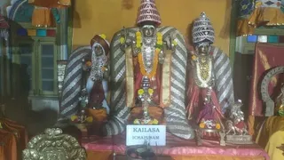 A photograph of a shrine displaying three seated deity murti in an ornate altar setting. The central figure wears a tall pointed crown with a striped ridged cone and is adorned with garlands of yellow and orange flowers and gold necklaces. Flanking figures wear similar crowns and garlands, with a smaller seated figure at the lower right. Food offerings and brass vessels sit before them on a pink-draped platform, and a red banner with a sign reading KAILASA ICHAPURAM is attached at the base. No people are visible in the frame.