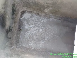 Photograph of a concrete water tank interior viewed from above, showing a shallow pool of murky grey water with ripples and foam on the surface. Dirty grey-brown concrete walls and a small round drain pipe opening are visible on the left edge. No people, deities or ritual objects are present; the scene shows the tank being cleaned or inspected as maintenance work.