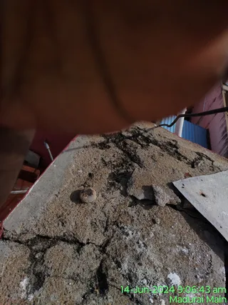 Close-up photograph of a cracked, grey concrete rooftop or tank top with a small rusted bolt or fitting near the centre and loose chipped debris. A blurred reddish-brown object. Beyond the parapet are a blue metal gate and a pink painted wall. A green timestamp in the lower right reads 14-Jun-2024 9:06:43 am and Madurai Main. No people are visible.