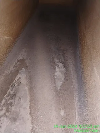 A photograph of the narrow interior of an empty concrete water tank or channel, with rough brown-beige walls and a damp, grey-brown floor streaked with pale residue. Light falls from the top of the frame and the far end recedes into shadow. No people, statues or ritual objects are visible. A green timestamp in the lower right reads 14-Jun-2024 9:37:22 am and Madurai Main.