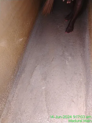 Photo of a narrow concrete channel or tank floor being cleaned. A person's bare legs and feet are visible at the top, and a straw broom with bristles is held at the upper edge of the frame. The floor is damp and grey with fine silt or residue, and a tan-coloured wall runs along the left side. A small stick and a scrap of white debris lie on the floor. A green timestamp in the bottom right reads 14-Jun-2024 9:17:03 am, Madurai Main.