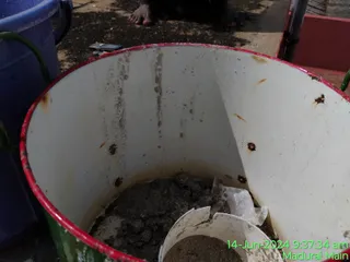 A close-up photo of the inside of a white metal bucket with a red rim and green handle, containing wet grey sludge and a broken white plastic piece. Rust spots mark the bucket's inner wall. A blue plastic bucket sits at the left edge, and bare feet of a person are partly visible at the top of the frame on a dusty ground surface. The bucket appears to be used in tank cleaning work. A timestamp in green reads 14-Jun-2024 9:37:34 am Madurai Main.
