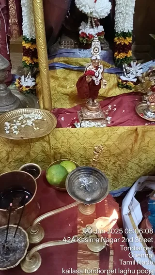 A small metal murti of a deity stands on a raised platform draped in gold and red cloth, decorated with a white flower garland and a tall ornate headdress with a round medallion, with a multi-coloured flower garland hanging on each side. Scattered white and red flower petals lie on the cloth and a brass plate of white flowers sits to the left. In the foreground are a copper vessel of water with incense sticks, a brass lamp with a lit flame, a bowl of green limes and a brass oil lamp with a small flame. The image carries a timestamp and a location caption reading kailasa tondiorpet laghu pooja. No people are visible.