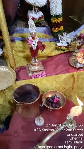 A small shrine scene photographed close up. At centre stands a metal deity murti in a red garment with a silver crown and a white-and-silver garland, set on a silver pedestal with pink flower petals scattered at its base. Behind it hang large garlands of white and yellow flowers. A copper vessel with smoke rising, a brass bowl of dark fruit or sweets with a pink flower, a lit oil lamp in a brass holder at lower right, and a brass tray with kumkum are arranged on gold and red cloths. No people are visible.