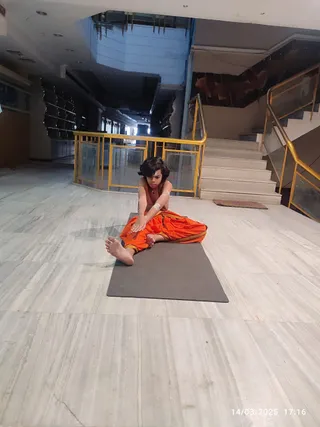 A photograph of a single child seated on a grey yoga mat on a pale marble floor in a large atrium with gold-framed glass railings and a marble staircase behind. The child, with dark shoulder-length hair and a vertical mark on the forehead, wears an orange-saffron dhoti-style garment with a gold border and a bare upper body with light markings on the arms. The child leans forward in a seated forward stretch, hands reaching toward the feet, bare feet extended. The timestamp 14/03/2025 17:16 appears in the lower right corner. a person's candidate status is not determinable from appearance, so the child is described by role only.