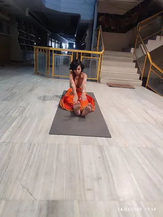 A photograph of a single person seated on a grey yoga mat on a polished marble floor, performing a forward-bend stretch with both legs extended and hands reaching toward the feet. The person has short dark hair, bare upper body with a rudraksha-style mala necklace, and wears an orange and yellow patterned dhoti-style lower garment. Behind them are a gold-railed balustrade, a staircase with white marble steps, and a corridor with glass panels. A date stamp in the lower right reads 14/03/2025 17:14.