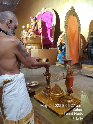 Photo inside a temple shrine. A shaved-headed priest with a grey beard and rudraksha mala, bare-chested and wearing a white dhoti with gold border, pours liquid from a brass vessel over a small gold-toned murti of a standing figure. Beside it stands a second gold-toned standing figure holding a cup, on a gold base with a metallic sri yantra-style meru piece in the middle. A brass plate with flower petals and a brass oil lamp sit nearby. Behind them is a gold seated deity murti garlanded with flowers under a magenta cloth, a blue-clad deity figure, a dark stone murti, and orange drapery in an arched niche. This appears to be an abhishekam being performed. The priest is a person, not a statue.