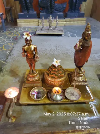 A photograph of a gilded ritual arrangement on a brass tray in an indoor temple hall. Two golden statues stand on either side: a small bare-chested young male figure with a plumeria flower on his head and hands in a gesture, and a taller female figure in an orange-red wrap holding a lamp-like object. In the centre is a golden multi-tiered sri yantra-style meru structure topped with a white plumeria flower. Before the statues sit brass bowls holding rudraksha beads with a flower, a small oil lamp burning, and an incense stick in a dish. A standing brass oil lamp burns at the left edge. In the background a silver deity murti group sits on a raised platform beneath dark stone statues, and the floor bears white and yellow kolam designs. No people are visible.