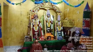 Interior shrine photo in a yellow-walled room decorated with blue and green paper streamers at the top. Three deity murtis stand behind a golden arch on a green-draped platform: the central figure wears a white garment and tall crown, while the flanking figures are garlanded with flower strings and wear colourful clothing. Gold staffs stand beside them, and brass lamps and offering vessels sit on the platform. A sign reads KAILASA TONDIARPET in gold lettering. On the right, a framed portrait of a man in a pink turban and dark garland is displayed beside a colourful decorative pillar. A timestamp in the corner reads 9 May 2025 04:39:59.