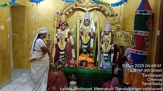 A woman in a white sari with an orange-bordered pallu and a white head covering stands with palms pressed together in namaste at the left of a shrine. Before her, three decorated murtis stand under a gold arch: each wears garlands of yellow, white and red flowers, and the central figure wears a tall silver crown. A small framed photo sits on an orange cloth at the base, with brass lamps and offerings in front. A sign reads KAILASA TONDIARPET BHARAT. Yellow walls, blue paper streamers and a striped cloth hanging at the right frame the room.