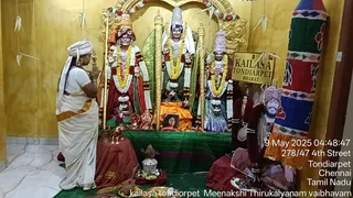A woman in a white sari with an orange-and-gold border stands with palms joined in namaskar before a shrine in a yellow-walled room. Three deity murtis stand on a green-draped altar, garlanded with yellow, white and red flower garlands, with a golden arch above. Brass lamps, a kalasha-like vessel and offerings sit on a red cloth in front. A sign reads KAILASA TONDIARPET BHARAT. At the right, a framed portrait labelled with a name shows a man with a white turban and a long beard; he is a portrait, not a physical person present. A colourful striped cloth hangs at the edge. Timestamp overlay reads 9 May 2025 04:48:47 with a Chennai address.