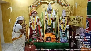 A photograph of a devotee woman in a white sari with an orange border and white head cloth standing at left with palms pressed together in namaskar before a shrine. The shrine holds three large painted deity murtis adorned with white and orange garlands: the central figure wears a silver crown and white garland, flanked by two figures in red and blue-green garments with silver crowns. Brass oil lamps and a small framed photograph sit on a green-draped altar, with a silver-faced Ganesha figure at the base. A sign reads KAILASA TONDIARPET BHARAT, and paper cranes hang from the yellow wall. Timestamp overlay reads 9 May 2025 04:49:02, 280 4th Street, Tondiarpet, Chennai, Tamil Nadu.