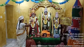 A woman in a white sari with an orange-and-gold border stands at left, wearing a white head-cloth, and extends her hand toward a shrine. The shrine holds three figures: a central deity murti in white and green with a tall crown and garland, flanked by two garlanded figures in red, green and white robes and crowns. Gold pillars and a gilded arch frame the shrine, with brass lamps, a green cloth and an orange-draped base below. A small framed photograph sits on the altar. Hanging paper decorations and blue and green garlands decorate the walls. At right a large cloth banner shows a man in an orange turban and robe, with a dated timestamp overlay.