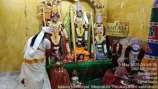A temple shrine room with yellow walls and gold-framed arches holds three deity murtis draped in green and red silk and heavy garlands of yellow and white flowers. A sign reads KAILASA TONDIARPET BHARAT. A devotee in white dhoti and shawl with a white head covering bends forward at left, facing the shrine, with an oil lamp and brass vessels on a red-draped table in front. At right, a framed photo and a large printed cutout portrait of a man with a saffron-maroon turban, rudraksha mala and garland stands beside the shrine, its face partly visible. A timestamp reads 9 May 2025 04:49:36, with address text 280 4th Street, Tondiarpet, Chennai, Tamil Nadu. Bottom caption reads kailasa tondiorpet Meenakshi Thirukalyanam vaibhavam.