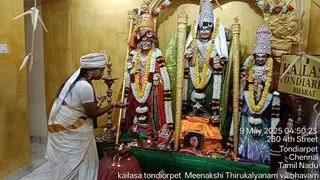 A photograph inside a temple shrine in Tondiarpet, Chennai, showing a priest in a white dhoti and white shawl with an orange border waving a flame before three deity murtis adorned with flower garlands. The central figure wears a crown and red garments, the right figure wears a tall ornamented crown, and a framed photo of a smiling woman rests on an orange cloth at the base. Brass oil lamps, a kalasha and offerings sit on a green-draped platform. A sign reads KAILASA TONDIARPET BHARAT, and a timestamp reads 9 May 2025 04:50:23.