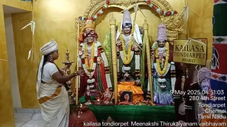 A photograph of a temple shrine with a golden-yellow wall. A woman in a white sari with an orange border and a white head wrap stands at left, holding a brass oil lamp (aarti) toward the deities. Three garlanded murti figures stand on a green-draped altar: at left a figure in a red and gold crown and red garments, at center a figure in white and gold with a tall crown, and at right a figure in a silver crown and blue-green garments. Marigold and white flower garlands adorn them. Offerings and a small framed portrait sit on the altar, with a KAILASA TONDIARPET BHARAT signboard at right. Some small paper cranes hang from the ceiling. A printed photo of a man in a turban appears at right edge. A timestamp reads 9 May 2025 04:51:03 with an address line.