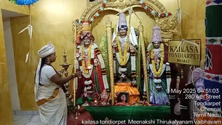 A photograph inside a golden-walled shrine room at KAILASA Tondiarpet. A woman in a white sari with an orange border, her head covered in white, holds an oil lamp toward a central group of deity murtis. The murtis stand on a green-draped altar, with a tall gold arch behind them, garlands of marigold and red flowers, and a small Ganesha murti at the base. A framed portrait of a man sits on an orange cloth at the foot of the altar. Brass oil lamps stand on either side, and a sign reads KAILASA TONDIARPET BHARAT. Hanging paper decorations are visible. A timestamp reads 9 May 2025 and the address 280 4th Street, Tondiarpet, Chennai, Tamil Nadu.