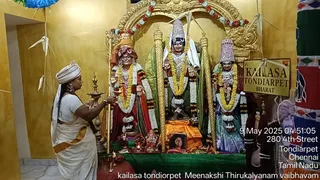 A woman in a white sari with an orange border and a white head cloth stands at left, holding a lit oil lamp in both hands toward a shrine of three deity murtis decorated with garlands of white and yellow flowers. The central figure is a standing deity with a tall crown and a gold-trimmed green garment; the figures at left and right wear red and teal garments with floral garlands. A small Ganesha murti and brass lamps sit on a green cloth in front. At lower centre is a framed photograph of a smiling woman in a dark outfit. A gold signboard at right reads KAILASA TONDIARPET BHARAT, and the shrine is set against a gold-painted wall.