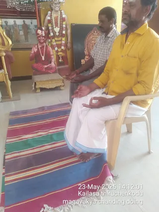 Two men sit on plastic chairs on a striped multicoloured rug in a room with yellow walls, meditating with hands in mudra and eyes closed. The man in front wears a yellow shirt and white dhoti with blue border, and the man behind wears a checked shirt. Behind them stand a life-size printed cutout of a garlanded figure in saffron robes and a flowered red headdress, plus a seated figure in red robes with a rudraksha mala and raised hand, both on a gold-edged display. A framed black-and-white group photo hangs at left and a bronze deity murti is at right. A timestamp reads 23-May-2025 14:12:01.