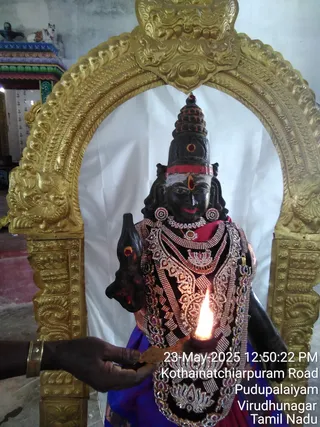 A photograph of a black stone deity murti standing within an ornate gold-toned arched frame against a white cloth backdrop. The murti wears a tall conical crown with red bindi marks, a red band across the forehead, white vibhuti, large earrings, layered silver-toned and jewelled necklaces, and a blue-and-red garment, and holds a trident-like staff. A lit oil lamp (deepa) is held before the figure by a hand entering from the lower left, with a flame burning in front of the chest. A timestamp and location caption are overlaid at the bottom right.